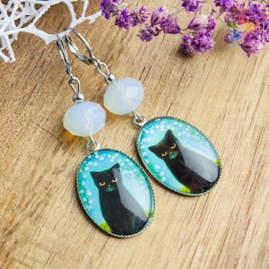 Può includere: Un paio di orecchini d'argento con un'immagine di un gatto nero su sfondo blu e una perla di opale bianca. Il gatto ha gli occhi gialli ed è circondato da fiori bianchi.