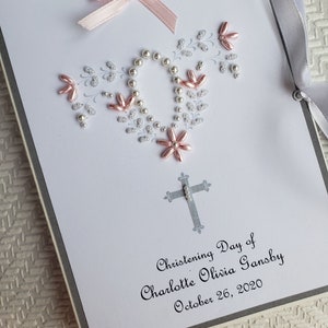 May include: A white card with a gray border and a pink bow. The card is decorated with white and pink beads in the shape of a wreath. The text on the card reads "Christening Day of Charlotte Olivia Gansby October 26, 2020".
