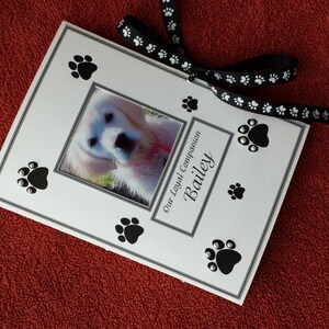 May include: A white card with a black border and paw print designs. A photo of a white dog is in the center of the card. The text "Our Loyal Companion Bailey" is printed on the card.