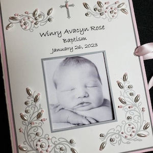 Peut inclure: Une carte de bapt&ecirc;me blanche avec une bordure rose. La carte pr&eacute;sente une croix, une photo d'un b&eacute;b&eacute; et le texte "Winry Avacyn Rose Bapt&ecirc;me 26 janvier 2023".
