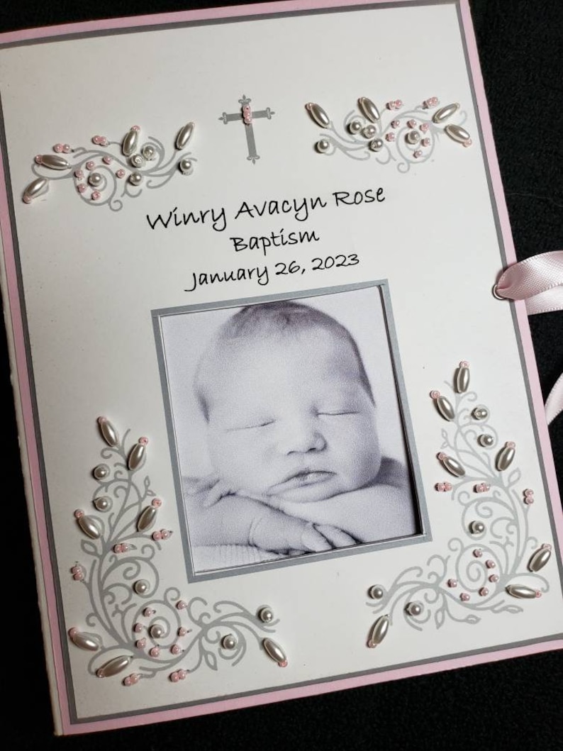 Peut inclure: Une carte de bapt&ecirc;me blanche avec une bordure rose. La carte pr&eacute;sente une croix, une photo d'un b&eacute;b&eacute; et le texte "Winry Avacyn Rose Bapt&ecirc;me 26 janvier 2023".