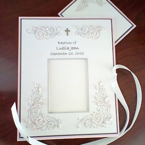 Peut inclure: Album photo blanc avec une bordure rose et une croix argent&eacute;e. L'album pr&eacute;sente un motif floral argent&eacute; et le texte "Baptism of Luella Jean September 25, 2022".