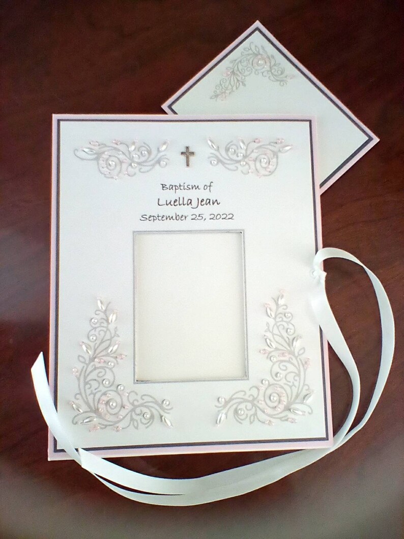 Peut inclure: Album photo blanc avec une bordure rose et une croix argent&eacute;e. L'album pr&eacute;sente un motif floral argent&eacute; et le texte "Baptism of Luella Jean September 25, 2022".