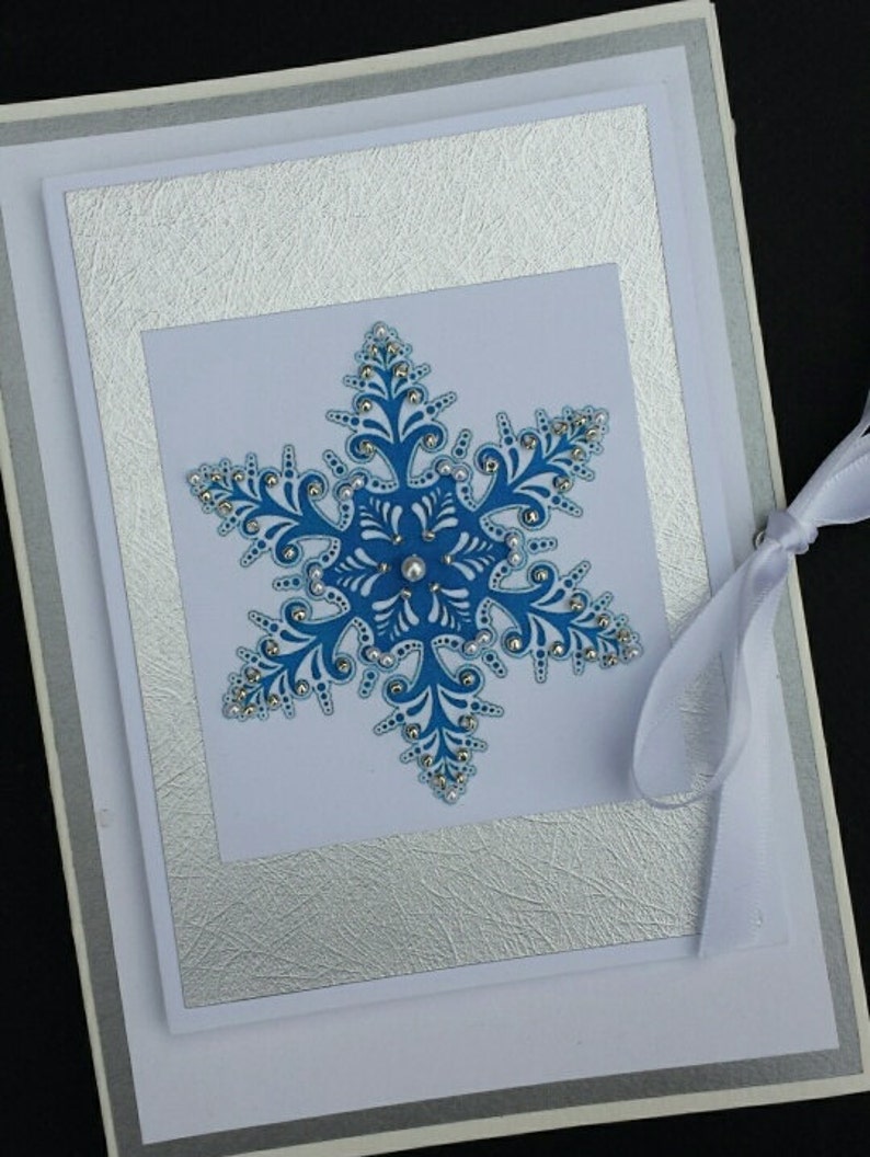 May include: A white card with a silver border and a blue snowflake in the center. The snowflake is decorated with white and silver glitter. The card has a white ribbon attached to it.