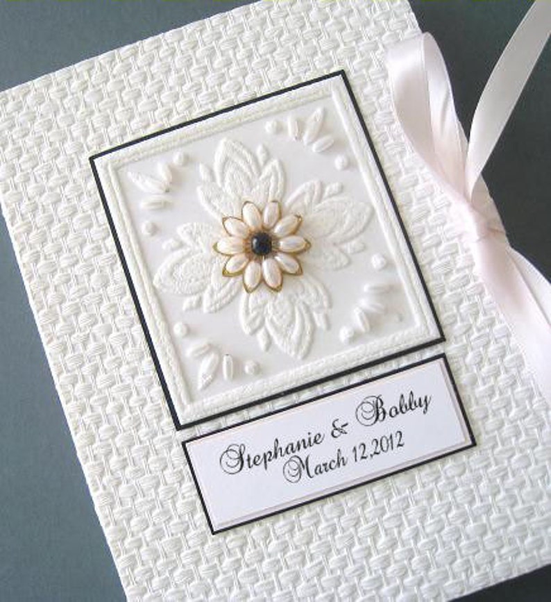 May include: White wedding guest book with a white embossed floral design and a black and white label with the text "Stephanie & Bobby March 12, 2012".