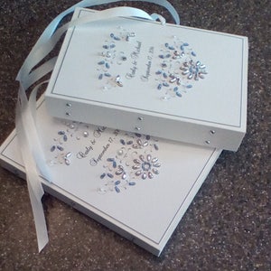May include: Two white wedding guest books with blue and silver embellishments. The top book has the text "Carly & Michael September 17, 2016". The bottom book has the text "Carly & Mic September 17, 201".