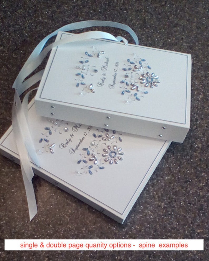 May include: Two white wedding guest books with blue and silver embellishments. The top book has the text "Carly & Michael September 17, 2016". The bottom book has the text "Carly & Mic September 17, 201".
