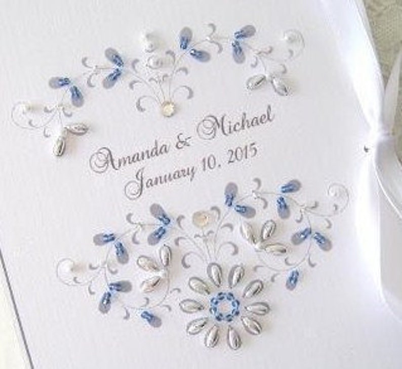 May include: A white wedding guest book with a silver and blue beaded design. The book is personalized with the names "Amanda & Michael" and the date "January 10, 2015".