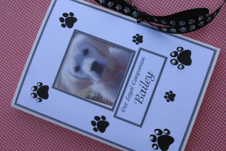 May include: A white photo frame with a black border and a picture of a white dog. The frame has black paw prints and silver glitter around the paw prints. The text "Our Loyal Companion Bailey" is printed on the frame.