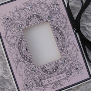 May include: A pink and black decorative frame with a cutout for a photo. The frame is adorned with intricate floral designs and rhinestones. The text "John & Heidi" is printed on the bottom of the frame.