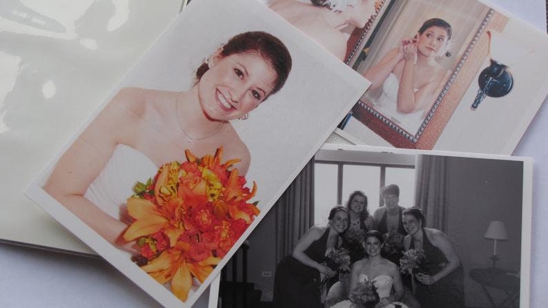 May include: A bride in a white dress holds a bouquet of orange and yellow flowers. The bouquet is made up of lilies, roses, and other flowers. The bride is smiling and looking at the camera.