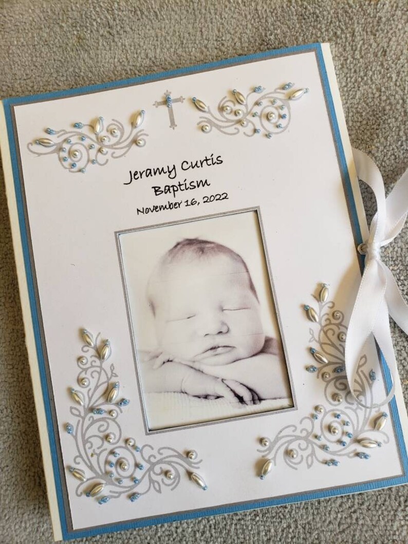 Peut inclure: Un album photo blanc avec une bordure bleue et des embellissements argent&eacute;s. L'album contient une photo d'un b&eacute;b&eacute; endormi au centre et le texte "Jeramy Curtis Baptism November 16, 2022" en haut.