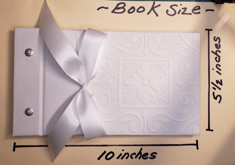 K&ouml;nnte beinhalten: Wei&szlig;es, gepr&auml;gtes Fotoalbum mit einer wei&szlig;en Satin Schleife. Das Album misst 25,4 cm in der Breite und 14 cm in der H&ouml;he. "Book Size" steht oben im Bild.