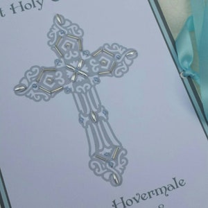 May include: A white card with a blue border and a silver cross with white and blue embellishments. The card reads "First Holy Communion" and "Hudson Hovermale April 14, 2018".