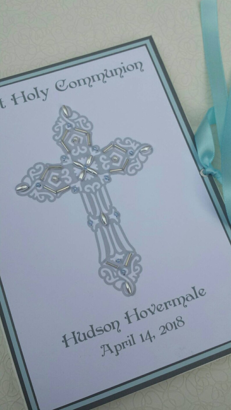 May include: A white card with a blue border and a silver cross with white and blue embellishments. The card reads "First Holy Communion" and "Hudson Hovermale April 14, 2018".