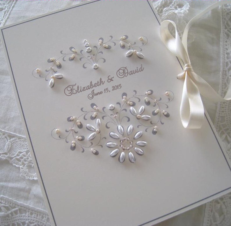 May include: A white wedding guest book with a gray floral design and pearl embellishments. The book is tied with a white satin ribbon and the text "Elizabeth & David June 15, 2015" is printed on the front.