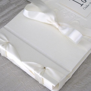 K&ouml;nnte beinhalten: Wei&szlig;es Hochzeitsalbum mit einem Satinband, das darum gebunden ist. Das Band ist auf der Vorderseite zu einer Schleife gebunden. Das Album hat ein wei&szlig;es, gepr&auml;gtes Design auf der Vorderseite. Der Text "October 12, 2017" ist auf einem kleinen wei&szlig;en Etikett gedruckt, das an der Vorderseite befestigt ist.