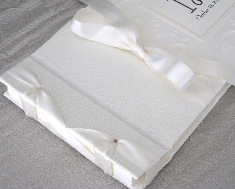 K&ouml;nnte beinhalten: Wei&szlig;es Hochzeitsalbum mit einem Satinband, das darum gebunden ist. Das Band ist auf der Vorderseite zu einer Schleife gebunden. Das Album hat ein wei&szlig;es, gepr&auml;gtes Design auf der Vorderseite. Der Text "October 12, 2017" ist auf einem kleinen wei&szlig;en Etikett gedruckt, das an der Vorderseite befestigt ist.