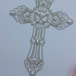 May include: A white card with a silver cross decorated with beads and the text "Child of God". The card also includes the name "Analiese Katherine Seeger" and the date "July 17, 2017".