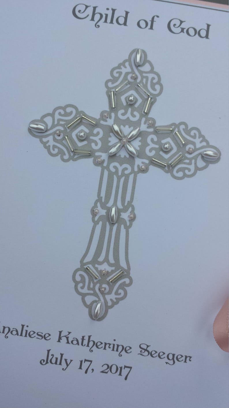 May include: A white card with a silver cross decorated with beads and the text "Child of God". The card also includes the name "Analiese Katherine Seeger" and the date "July 17, 2017".