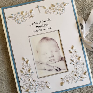 Peut inclure: Un album photo blanc avec une bordure bleue et des embellissements argent&eacute;s et bleus. L'album contient une photo d'un b&eacute;b&eacute; endormi. Le texte sur l'album est "Jeramy Curtis Bapt&ecirc;me 16 novembre 2022".
