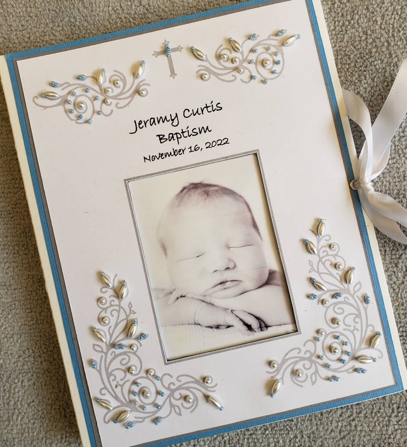 Peut inclure: Un album photo blanc avec une bordure bleue et des embellissements argent&eacute;s et bleus. L'album contient une photo d'un b&eacute;b&eacute; endormi. Le texte sur l'album est "Jeramy Curtis Bapt&ecirc;me 16 novembre 2022".