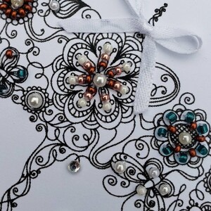 May include: A white card with a black and white intricate design featuring a butterfly, flowers, and a white ribbon bow. The design is embellished with copper, white, and teal beads.