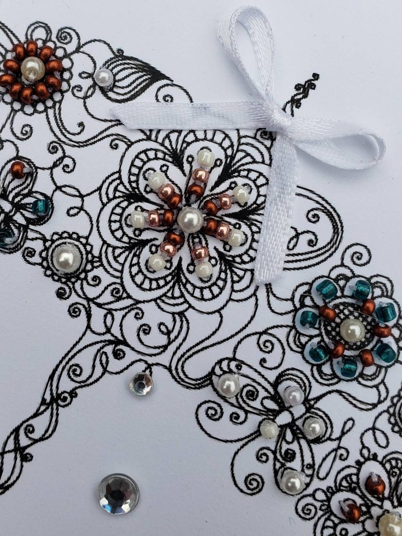 May include: A white card with a black and white intricate design featuring a butterfly, flowers, and a white ribbon bow. The design is embellished with copper, white, and teal beads.