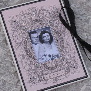 May include: A pink wedding scrapbook with a black ribbon closure. The cover features a decorative floral design and a framed photo of a couple. The text "John & Heidi" is printed on the cover.