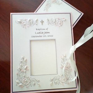 Peut inclure: Un album photo blanc avec une bordure rose et une croix argent&eacute;e. L'album est orn&eacute; d'un motif floral argent&eacute; et rose et d'un cadre photo avec le texte "Baptism of Luella Jean September 25, 2022".