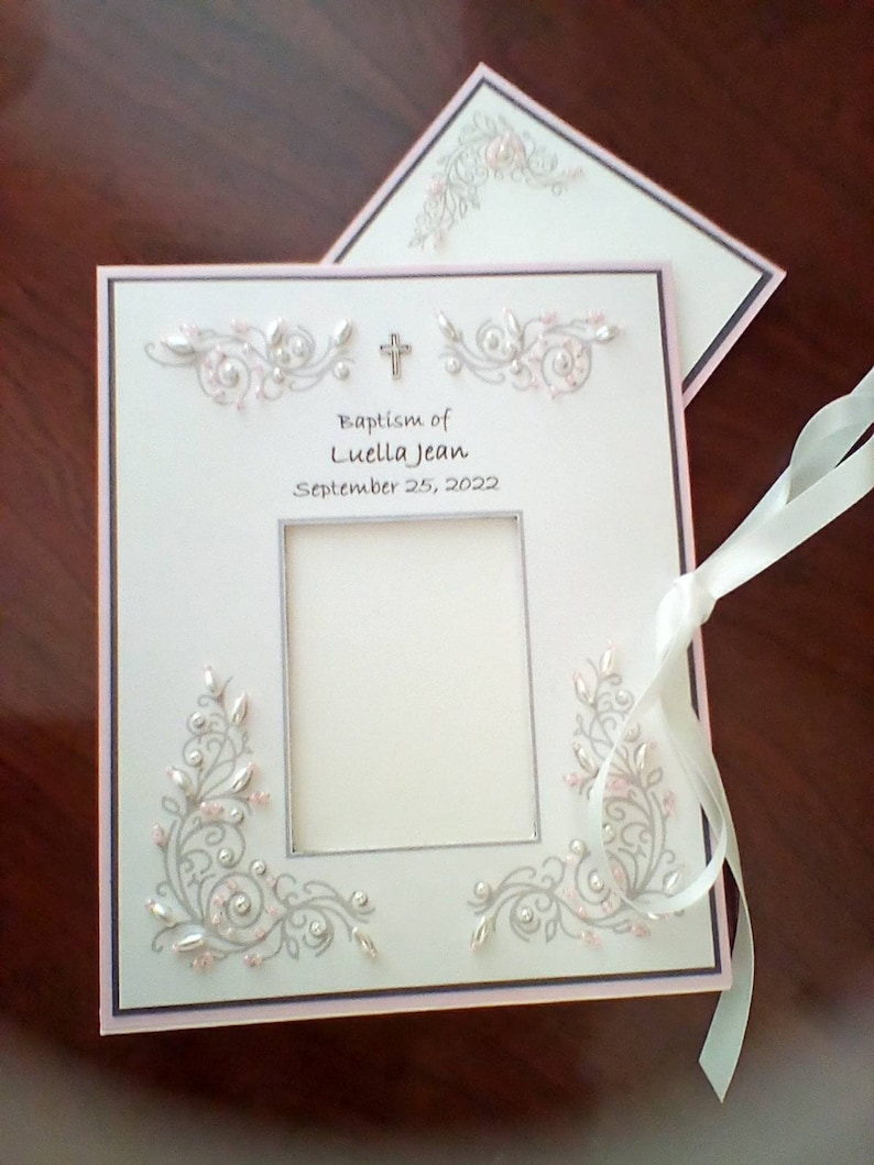 Peut inclure: Un album photo blanc avec une bordure rose et une croix argent&eacute;e. L'album est orn&eacute; d'un motif floral argent&eacute; et rose et d'un cadre photo avec le texte "Baptism of Luella Jean September 25, 2022".