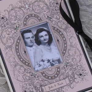 May include: A vintage-style wedding album with a black ribbon closure. The cover features a decorative frame with a black and white photo of a bride and groom. The text "John & Heidi" is printed on the bottom of the album.