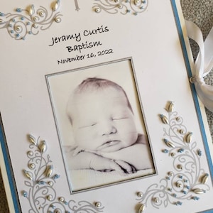 Peut inclure: Un album photo blanc avec une bordure bleue et des embellissements argent&eacute;s. La couverture pr&eacute;sente une photo d'un b&eacute;b&eacute; endormi, une croix et le texte "Jeramy Curtis Bapt&ecirc;me 16 novembre 2022".