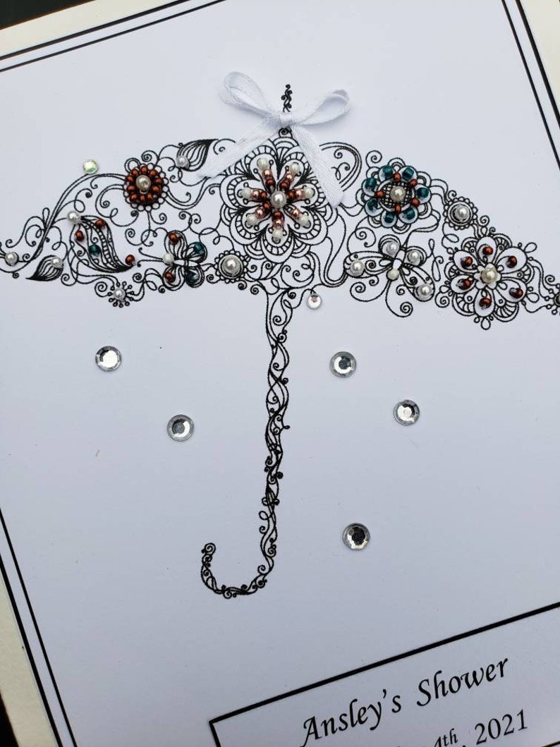 May include: A white card with a black and white illustration of an umbrella with a white bow. The umbrella is decorated with pearls and rhinestones. The card has the text "Ansley's Shower 1th 2021" at the bottom.