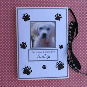 May include: A white photo album with a black ribbon and paw print designs. The album has a photo of a white dog with black eyes and a black nose. The text "Our Loyal Companion Bailey" is printed on the front of the album.
