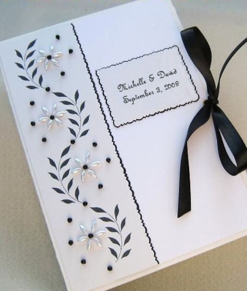May include: A white wedding guest book with black trim and silver flowers. The book has a black ribbon tied around it. The book has a label that reads 'Michelle & David September 3, 2009'.