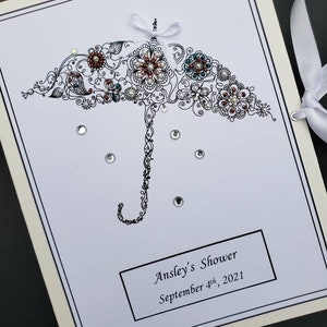 May include: A white card with a black and white illustration of an umbrella with flowers and rhinestones. The text "Ansley's Shower September 4th, 2021" is printed on the card.