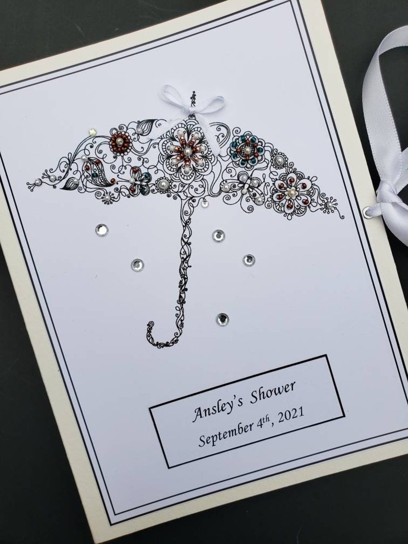 May include: A white card with a black and white illustration of an umbrella with flowers and rhinestones. The text "Ansley's Shower September 4th, 2021" is printed on the card.
