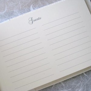 K&ouml;nnte beinhalten: Ein wei&szlig;es G&auml;stebuch mit einem schlichten, eleganten Design. Der Einband hat eine dezente Textur und das Wort "Guests" ist in einer kursivschriftlichen Schriftart gedruckt.