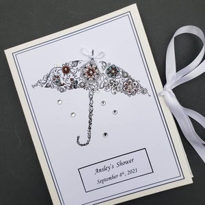 May include: A white card with a black and white illustration of an umbrella with rhinestones. The umbrella is decorated with flowers and swirls. The text "Ansley's Shower September 4th, 2021" is printed on the card.