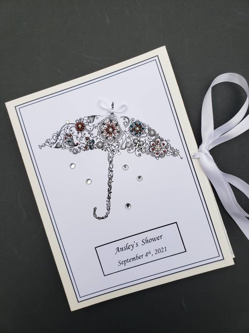 May include: A white card with a black and white illustration of an umbrella with rhinestones. The umbrella is decorated with flowers and swirls. The text "Ansley's Shower September 4th, 2021" is printed on the card.