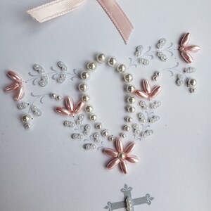 May include: A white card with a pink satin ribbon bow at the top. The card is decorated with white and pink beads in the shape of a cross and flowers. The cross is made of gray glitter.