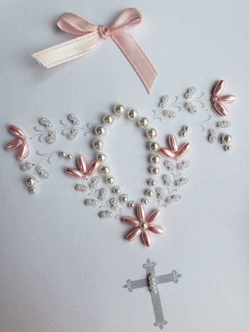May include: A white card with a pink satin ribbon bow at the top. The card is decorated with white and pink beads in the shape of a cross and flowers. The cross is made of gray glitter.