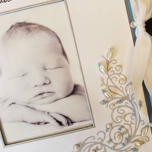 Peut inclure: Un album photo blanc avec un cadre argent&eacute; et une photo en noir et blanc d'un b&eacute;b&eacute; &agrave; l'int&eacute;rieur. L'album a un design d&eacute;coratif bleu et argent avec des perles argent&eacute;es et bleues.
