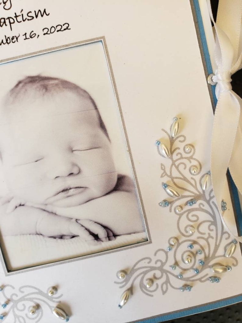 Peut inclure: Un album photo blanc avec un cadre argent&eacute; et une photo en noir et blanc d'un b&eacute;b&eacute; &agrave; l'int&eacute;rieur. L'album a un design d&eacute;coratif bleu et argent avec des perles argent&eacute;es et bleues.