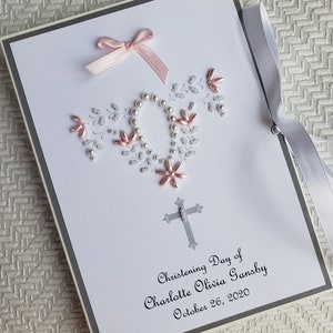 May include: A white card with a silver border and a pink ribbon bow. The card is decorated with white and pink beads in the shape of a cross. The text on the card reads "Christening Day of Charlotte Olivia Gansby October 26, 2020".