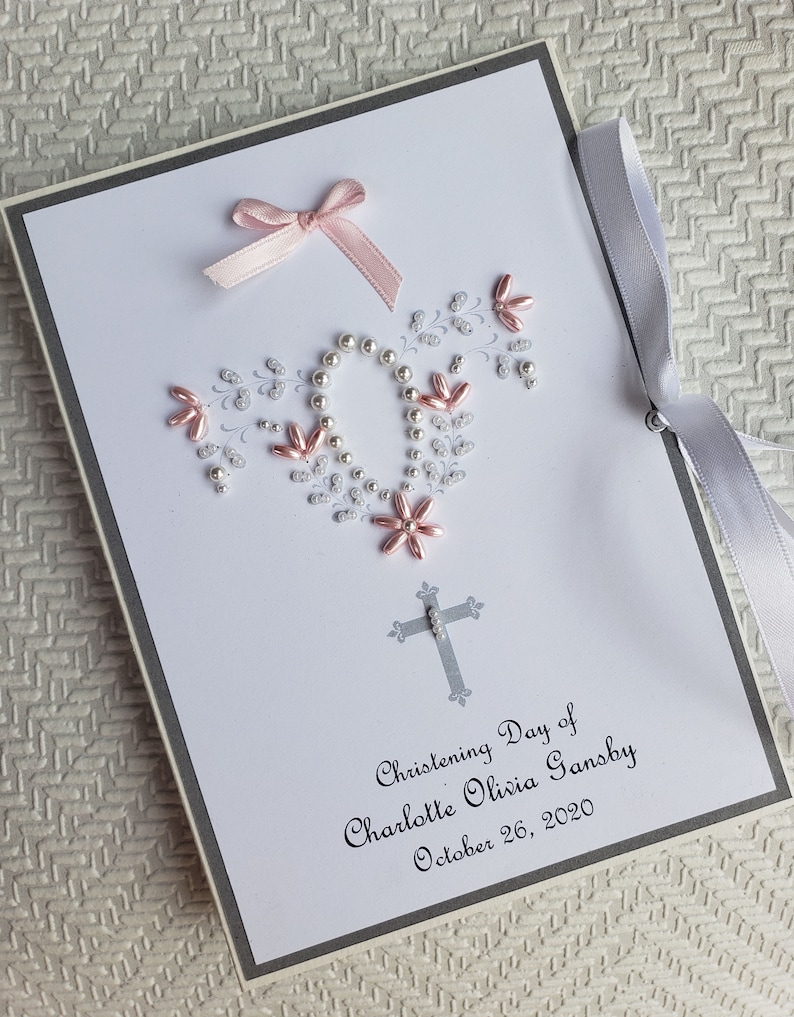 May include: A white card with a silver border and a pink ribbon bow. The card is decorated with white and pink beads in the shape of a cross. The text on the card reads "Christening Day of Charlotte Olivia Gansby October 26, 2020".