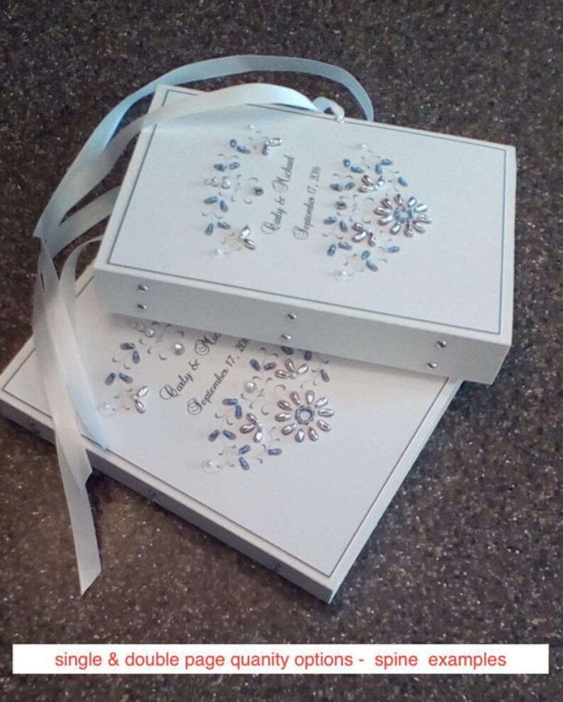 May include: Two white wedding guest books with blue and silver embellishments. The top book has the text "Carly & Michael September 17, 2018".
