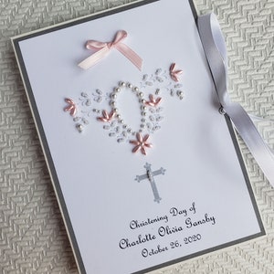 May include: A white card with a gray border and a pink bow. The card has a silver cross and a floral design made of silver and pink beads. The text on the card reads "Christening Day of Charlotte Olivia Gansby October 26, 2020".