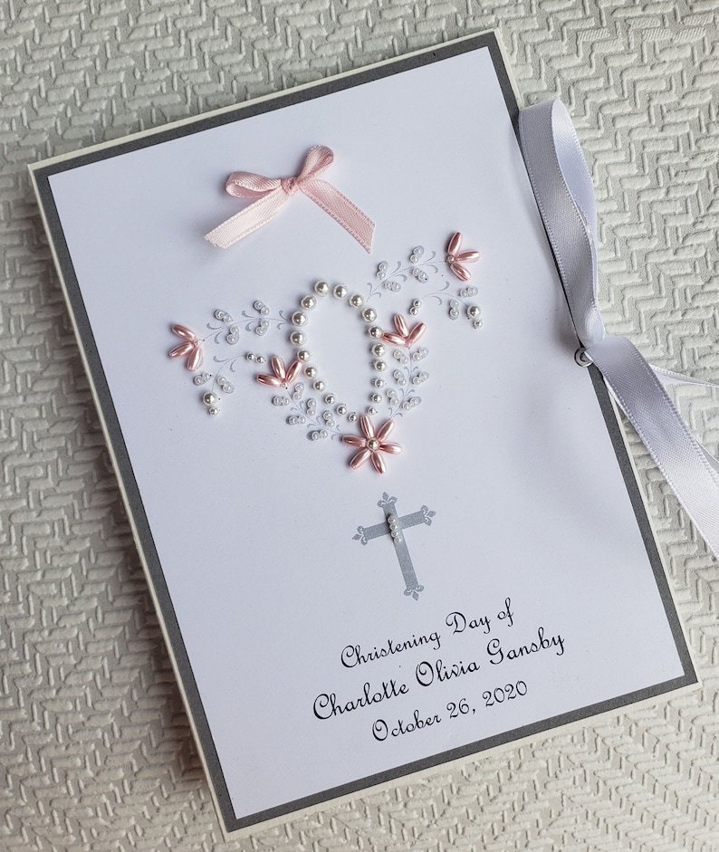 May include: A white card with a gray border and a pink bow. The card has a silver cross and a floral design made of silver and pink beads. The text on the card reads "Christening Day of Charlotte Olivia Gansby October 26, 2020".